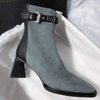 A profile shot of a single grey suede boot against a white tissue paper background, showing the contrast between the grey suede body and the black leather heel counter and ankle strap.