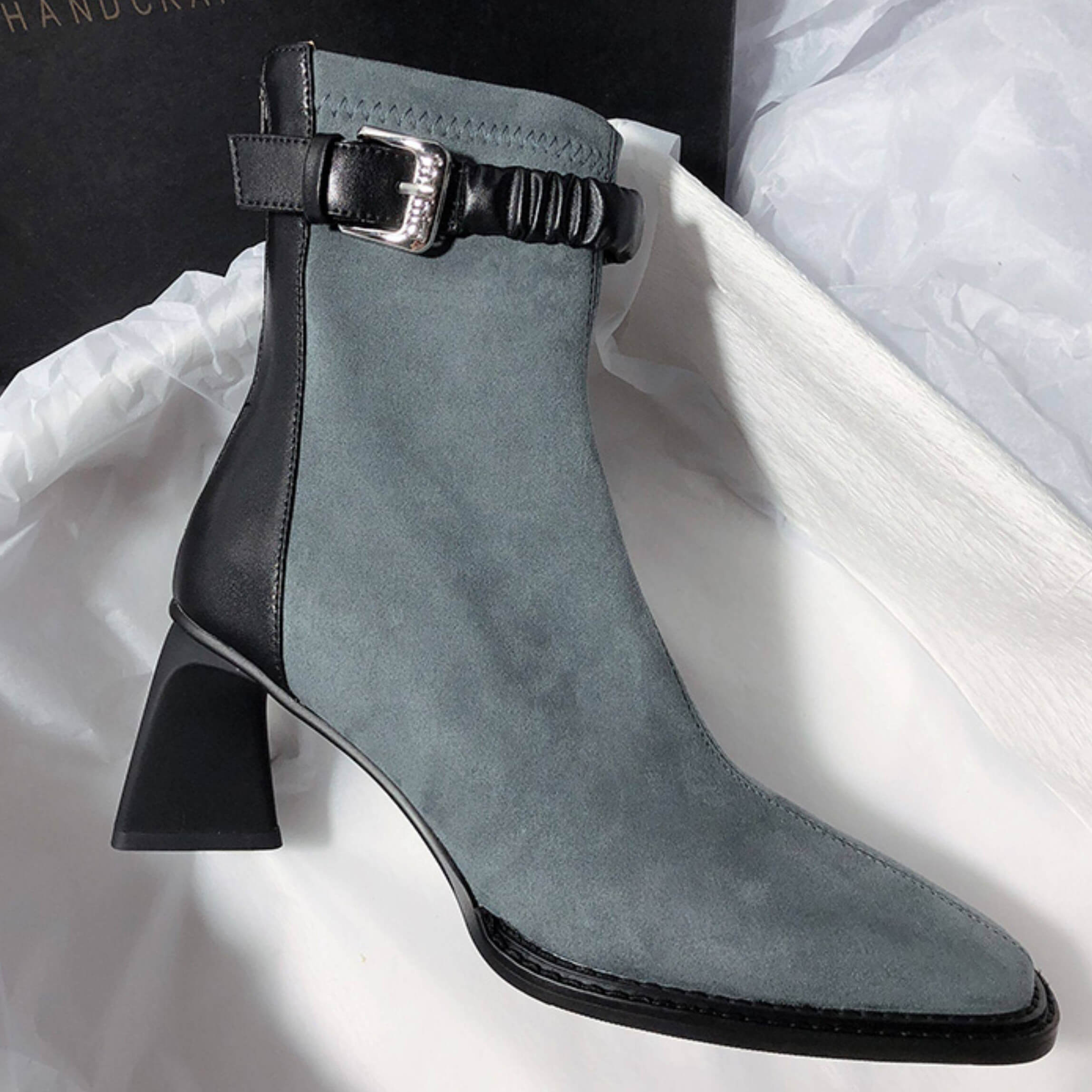 A profile shot of a single grey suede boot against a white tissue paper background, showing the contrast between the grey suede body and the black leather heel counter and ankle strap.