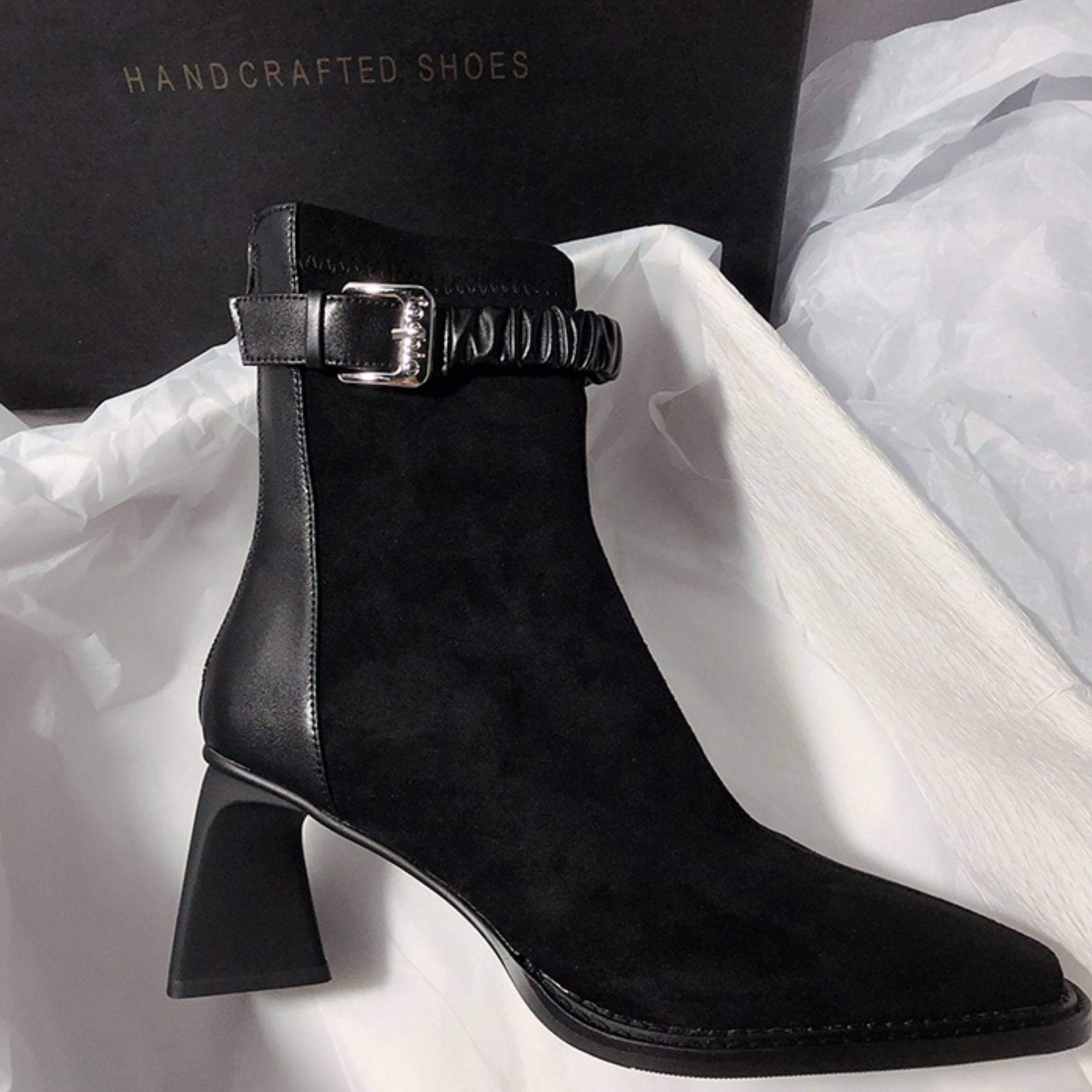 A profile shot of the black version of the ankle boot, showcasing the same buckled ruched strap and flared heel in an all-black colorway.