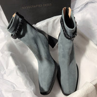 Two grey suede boots arranged inside a "HANDCRAFTED SHOES" box, one showing the side profile and the other showing the front view.
