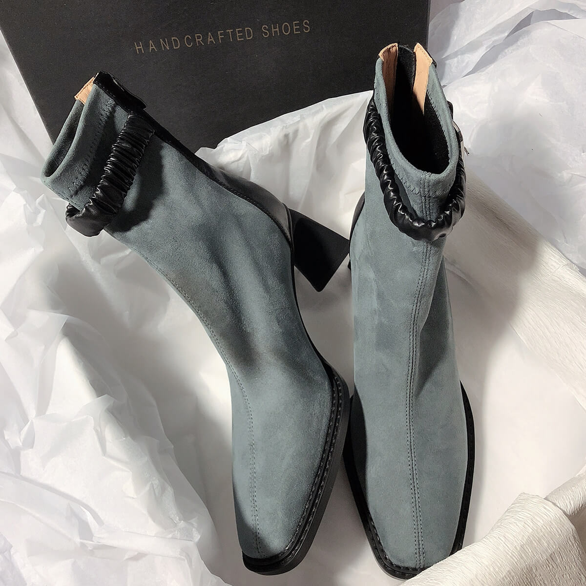 Two grey suede boots arranged inside a "HANDCRAFTED SHOES" box, one showing the side profile and the other showing the front view.