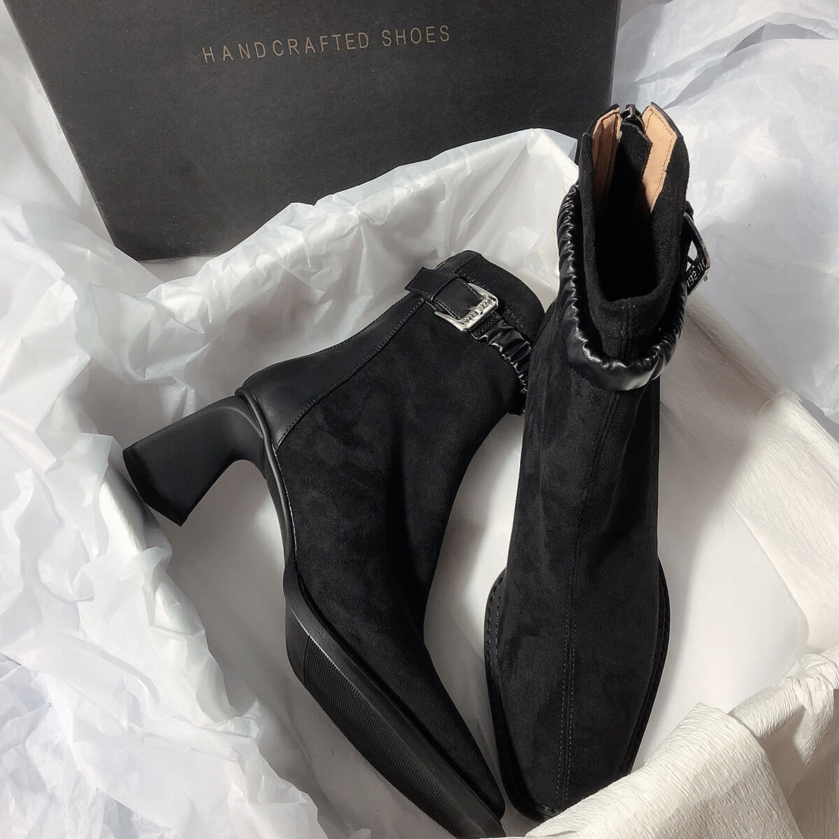 Two black suede boots arranged inside their packaging, echoing the layout of the grey pair to show the alternative color option.