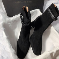 A pair of black suede ankle boots presented in a black "HANDCRAFTED SHOES" box with white tissue paper. The image displays the boots from two angles, highlighting the matching black ruched ankle strap and the structural black block heel.