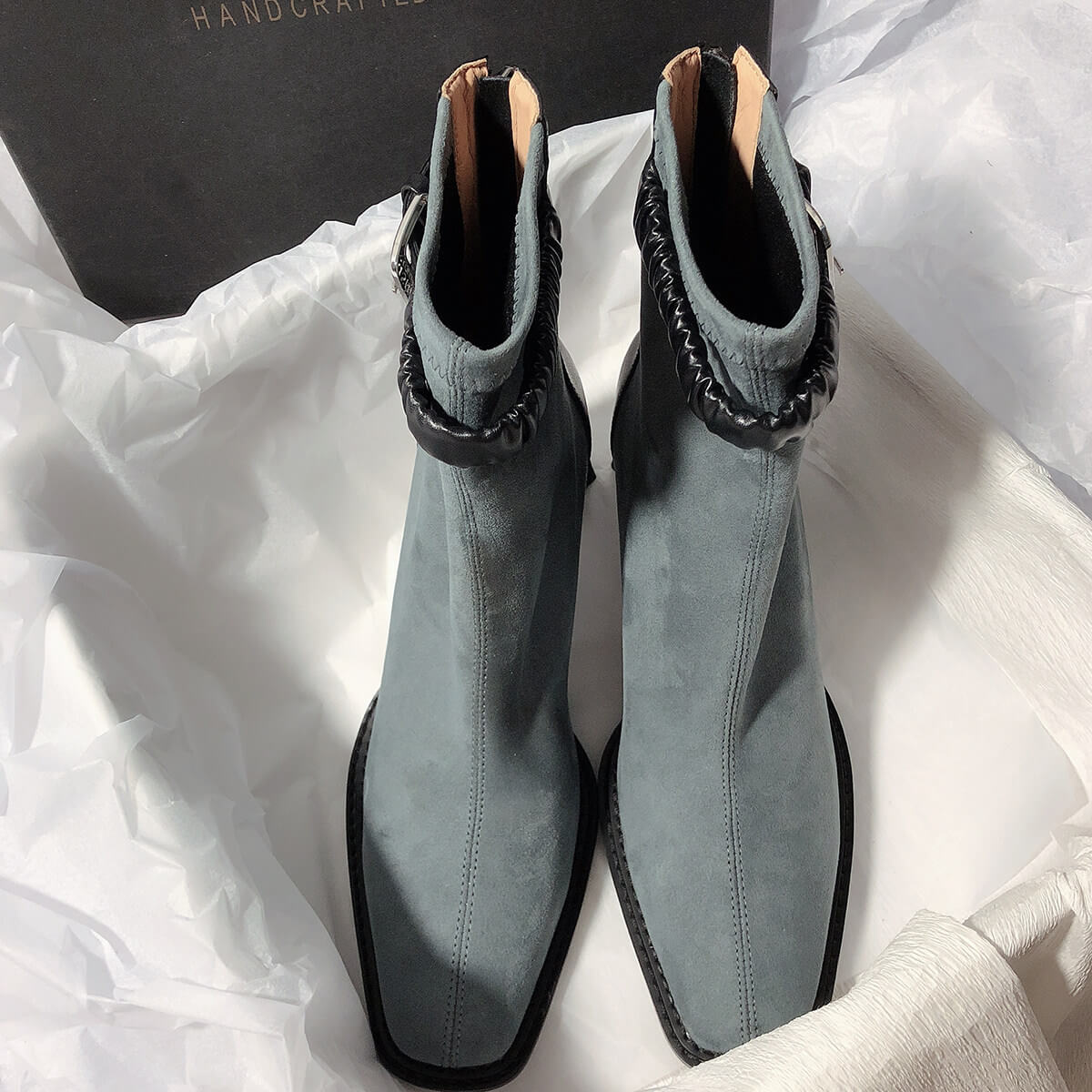 A top-down view of a pair of grey suede ankle boots sitting in a box lined with white tissue paper. The boots feature a distinctive black ruched leather strap around the ankle and a center seam running down the front.
