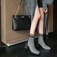 A full-length lifestyle shot of the grey ankle boots being worn. The outfit includes a long grey cardigan and a black quilted luxury handbag with a gold chain strap.

