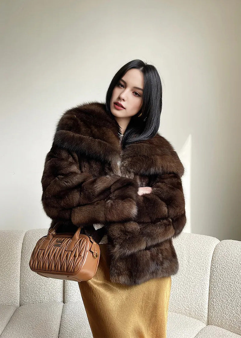 The woman is shown from the waist up, standing with her arms crossed. This pose emphasizes the thick, tiered construction of the fur sleeves and the large, fluffy collar of the jacket.