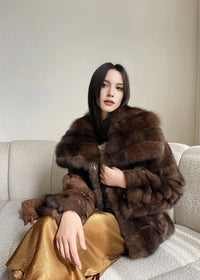 The woman is seated on a textured off-white curved sofa. She is wearing the dark brown fur coat and a gold skirt, leaning slightly forward with a calm expression.