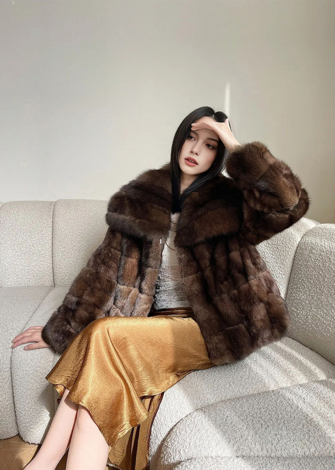  The woman is seated on the sofa, leaning back with one hand raised to her forehead as if shielding her eyes from the sun. The photo highlights the oversized fit of the fur jacket and the reflective texture of the gold skirt.