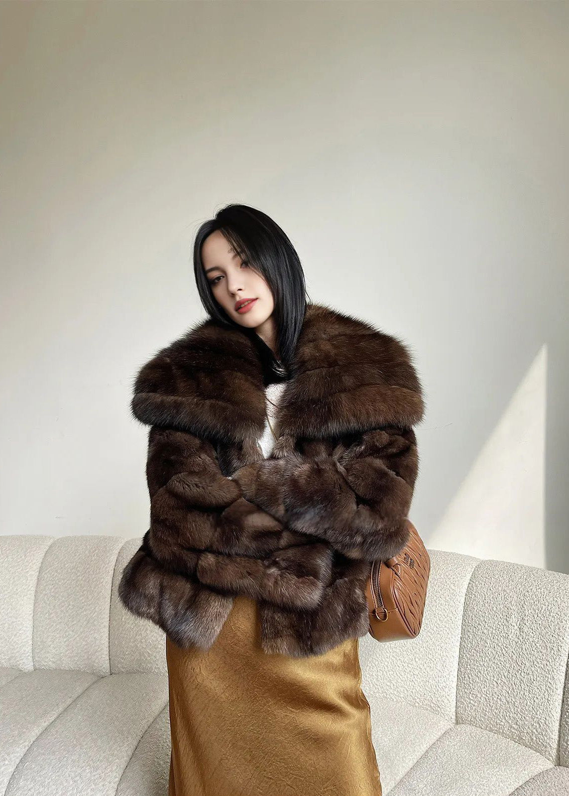 A standing shot where the woman has her arms tightly crossed over her chest, burying her hands in the thick brown fur. She looks toward the camera with a slight tilt of her head.