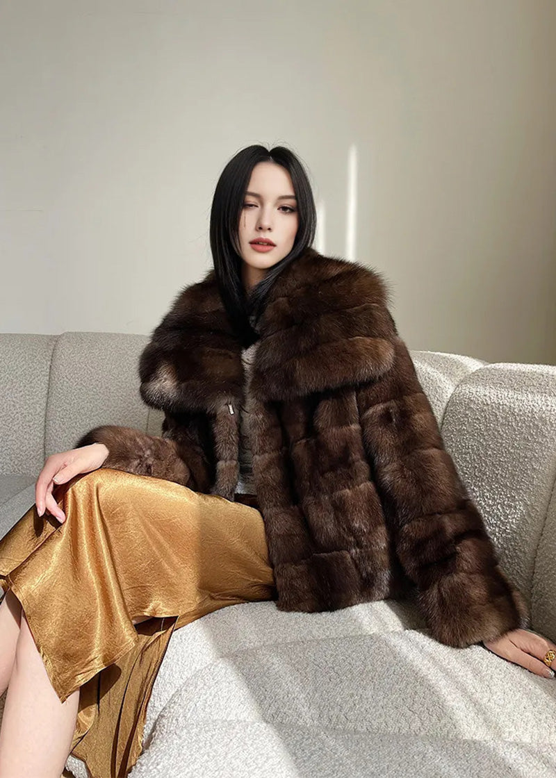 A wide-angle shot of the woman sitting on the white sofa, showcasing the full length of the gold satin skirt and the plush texture of the brown fur jacket under natural sunlight.