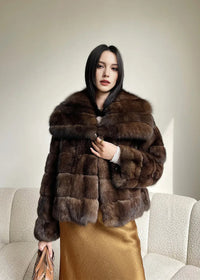 A front-facing portrait of the woman standing, wearing the brown fur jacket and gold skirt. She is holding the front of the jacket closed with her right hand and carrying the tan leather bag in her left.