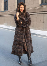A full-length photo of a woman posing with the hood of her thick brown fur coat pulled up, wearing black boots and standing on an empty asphalt road.