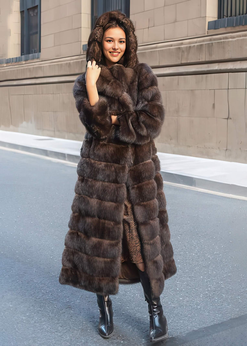 A full-length photo of a woman posing with the hood of her thick brown fur coat pulled up, wearing black boots and standing on an empty asphalt road.