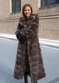 A woman standing on a city street, smiling warmly with her arms crossed. She is wearing a heavy, floor-length brown fur coat with distinct horizontal paneling.