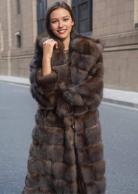 A medium shot of a smiling woman posing in a luxurious, textured brown fur coat. She has her hand near her collar and is standing on a paved street.