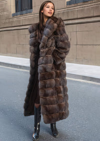 A woman looking off to the side with a slight smile, posing in an open, full-length brown fur coat that shows a dark dress and black boots.