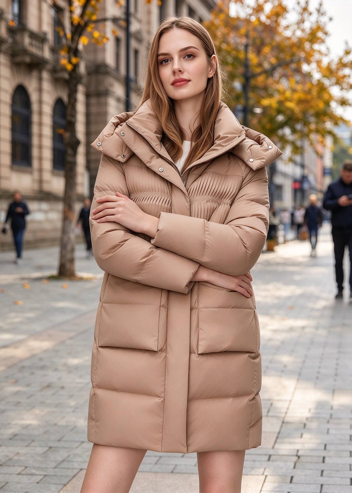 Front three-quarter angle of women's camel hooded puffer jacket featuring arms-crossed pose, pleated chest accents, and deep flap pockets.