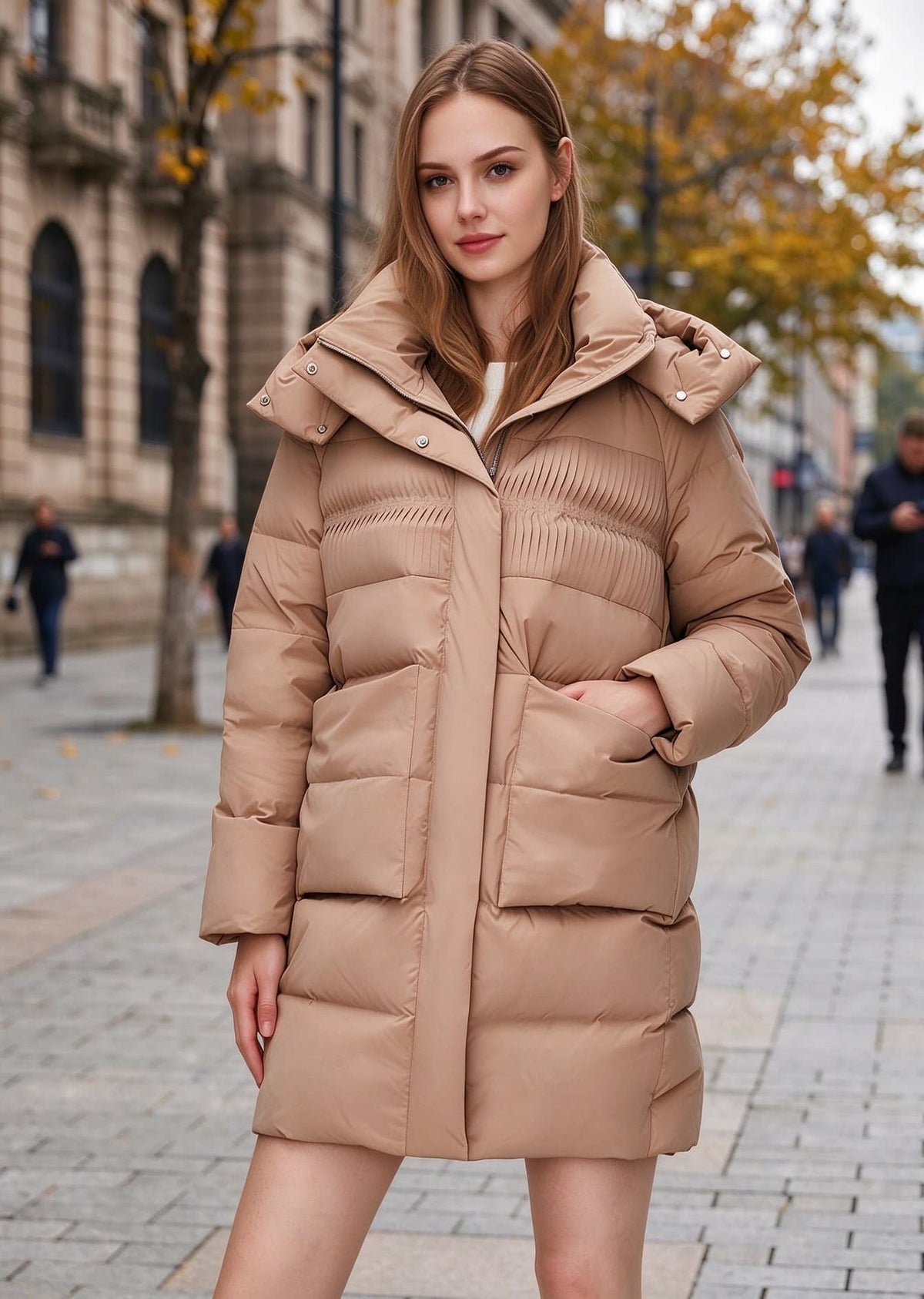 Front view of woman wearing a camel khaki mid-length hooded down puffer coat with pleated yoke details and large flap pockets on a city street.