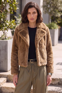 A woman wearing a brown faux fur jacket over a black top and olive green pants, standing outdoors in front of a modern building with stone steps and greenery.