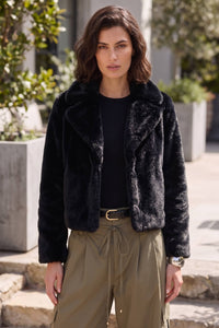 A woman wearing a black faux fur cropped jacket over a black top and olive green drawstring pants, standing outdoors in front of a modern building.