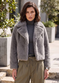 A woman wearing a gray fuzzy cropped jacket over a dark gray turtleneck, paired with olive green drawstring pants, standing outdoors in front of a modern building.