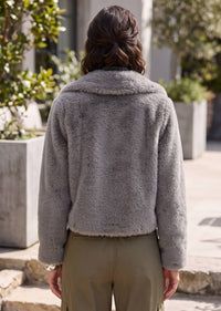 A person viewed from behind wearing a fuzzy light gray cropped jacket with a wide collar and olive green pants, standing outdoors in front of a building with planters.
