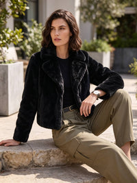 A woman sits outdoors on stone steps wearing a black fuzzy cropped jacket over a black top and olive green cargo pants.