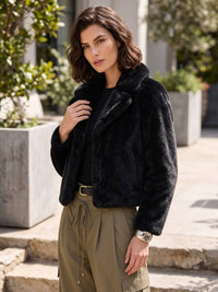 A woman wearing a black faux fur cropped jacket, olive green cargo pants, and a black top, standing outdoors in front of a modern building with stone steps and greenery.