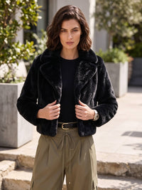 A woman wearing a black faux fur cropped jacket over a black top and olive green drawstring pants, standing outdoors in front of stone steps and greenery.