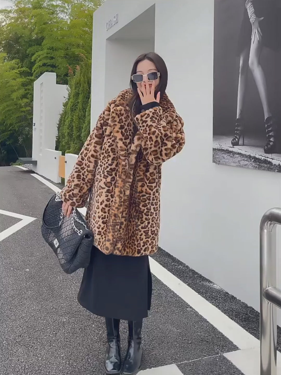 Model showcasing the Vivian Seven Luxury Oversized Leopard Faux Fur Coat. This long-sleeve leopard jacket features a plush texture and a relaxed, oversized fit. Styled with a black midi skirt, boots, and a quilted handbag for a high-end winter fashion look.
