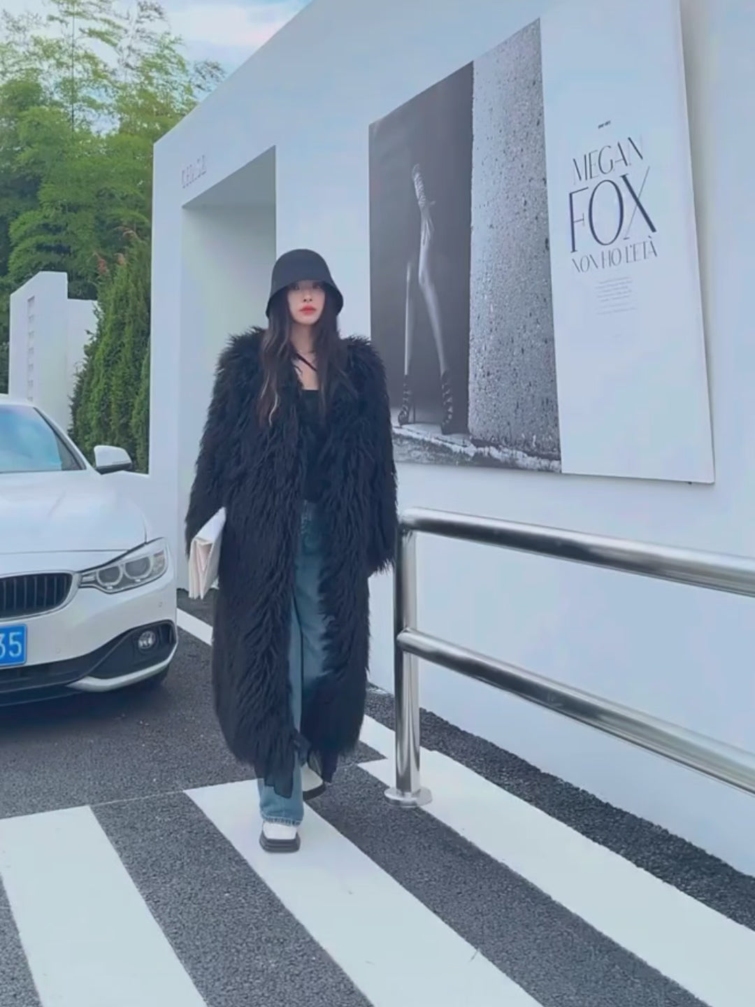 Model wearing the Vivian Seven Women's Longline Shaggy Black Faux Fur Coat with sheer ruffle trim. Styled with a black bucket hat, wide-leg blue jeans, and white platform loafers. A luxury winter outerwear piece for boho chic and streetwear styles.