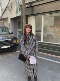 A young woman models a long Vivan Seven gray plaid wool-blend coat while walking and posing on a city street.