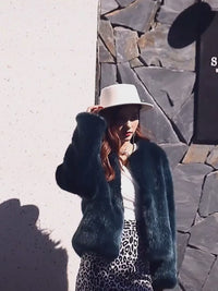 Model wearing the Vivian Seven Women's Plush Faux Fur Cropped Jacket in deep emerald green. Features a soft, silky faux mink texture and a collarless design. Styled with a white wide-brim hat, leopard print skirt, and layered jewelry for a glamorous winter outfit