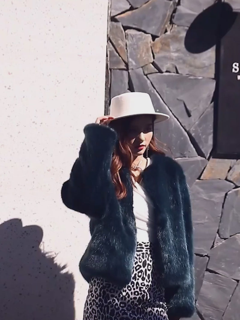 Model wearing the Vivian Seven Women's Plush Faux Fur Cropped Jacket in deep emerald green. Features a soft, silky faux mink texture and a collarless design. Styled with a white wide-brim hat, leopard print skirt, and layered jewelry for a glamorous winter outfit