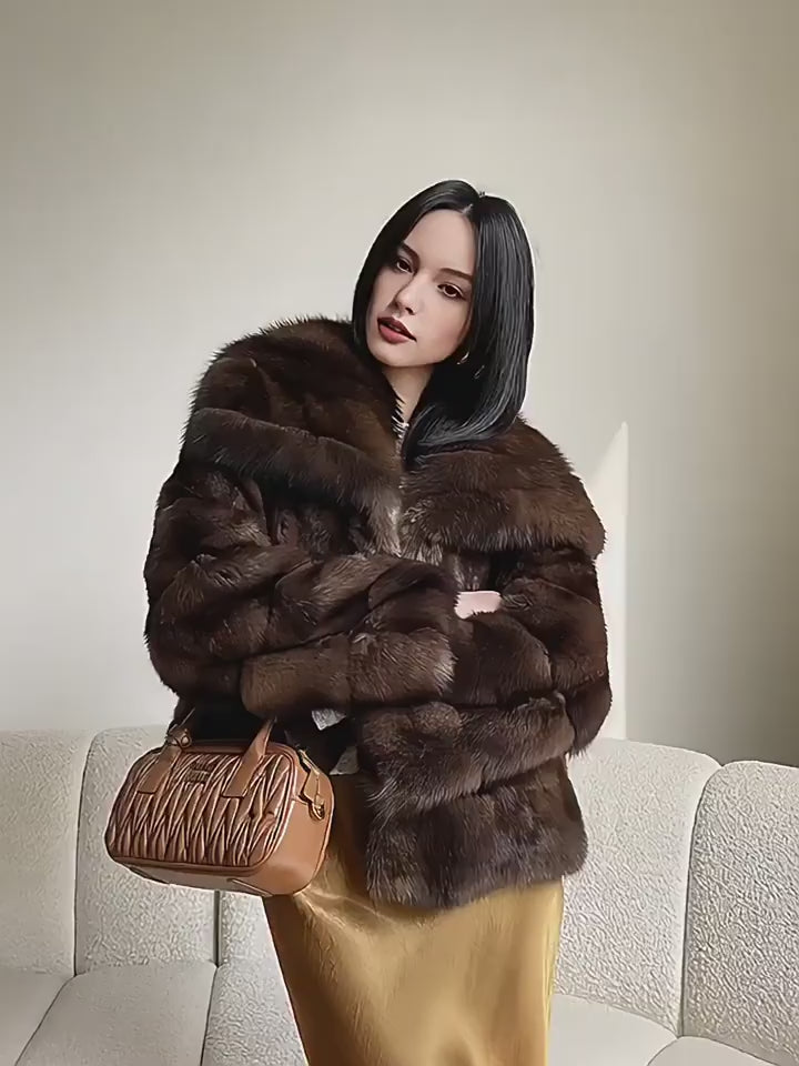 A woman models a luxurious, dark brown faux fur jacket with horizontal panelling and a wide, plush collar. She poses in an elegant indoor setting, pairing the jacket with a shimmering gold satin skirt and a quilted brown handbag. The video captures her standing and then gracefully sitting on a white sofa, highlighting the soft texture and high-fashion aesthetic of the outfit.