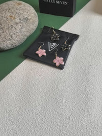 A close-up video showcasing four enamel flower drop earrings (two pink, two black with gold-toned trim) resting on a black VIVIAN SEVEN branded drawstring pouch. The video focuses on a hand picking up one of the pink earrings, turning it to display the front (pink enamel) and the back (gold-toned textured metal), before placing it back down. The background includes a grey rock and a black box.