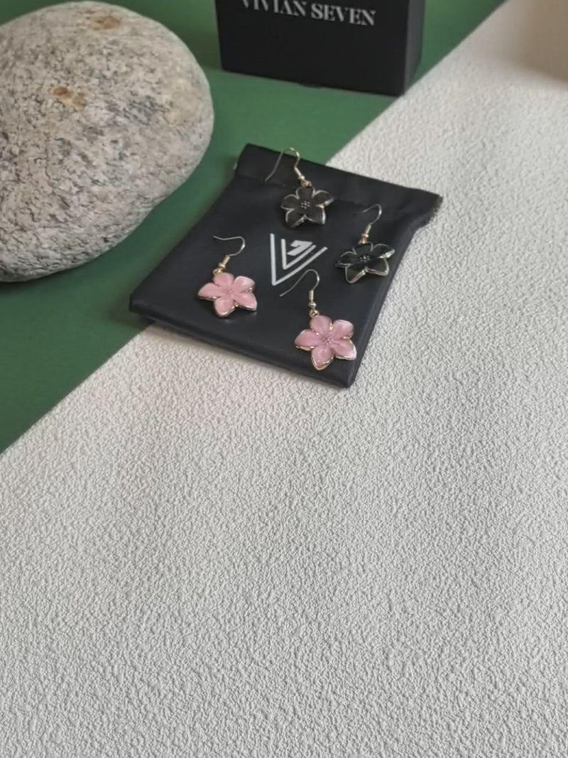 A close-up video showcasing four enamel flower drop earrings (two pink, two black with gold-toned trim) resting on a black VIVIAN SEVEN branded drawstring pouch. The video focuses on a hand picking up one of the pink earrings, turning it to display the front (pink enamel) and the back (gold-toned textured metal), before placing it back down. The background includes a grey rock and a black box.
