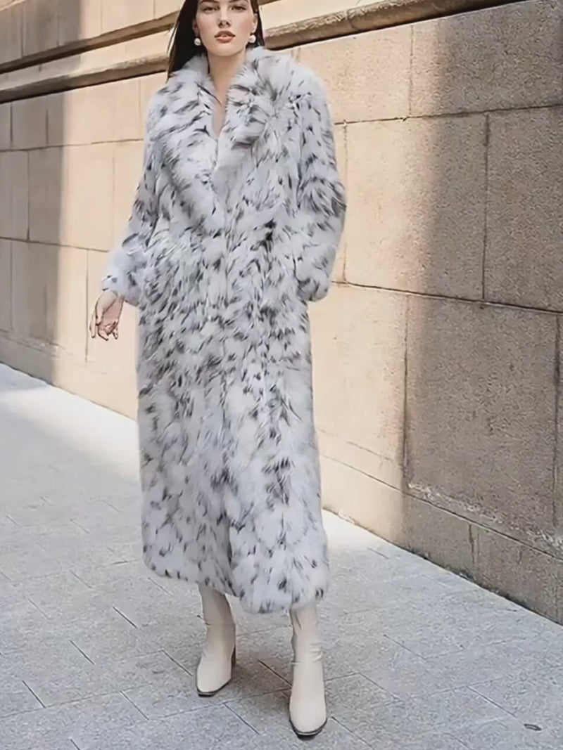 A woman models a luxurious, ankle-length faux fur coat with a white base and striking black spotted pattern. She walks confidently along a sunlit city sidewalk next to a stone building, showcasing the coat’s wide lapels and voluminous fit, paired with cream-colored ankle boots.