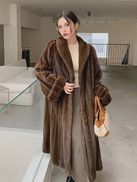A woman models two different luxurious faux fur coats in an elegant setting. The first is a long, rich brown coat with horizontal panels, paired with a beige top, dark trousers, and a tan-and-white handbag. The second is a sleek, dark mahogany coat worn over a white cardigan and long denim skirt. The video captures her posing and walking, emphasizing the plush texture and sophisticated draping of the outerwear.