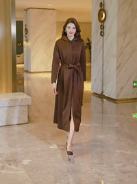 A product showcase video for a Vivian Seven chocolate brown hooded long trench coat. The video features a model walking through a modern interior, demonstrating the coat's tailored fit and fluid movement.